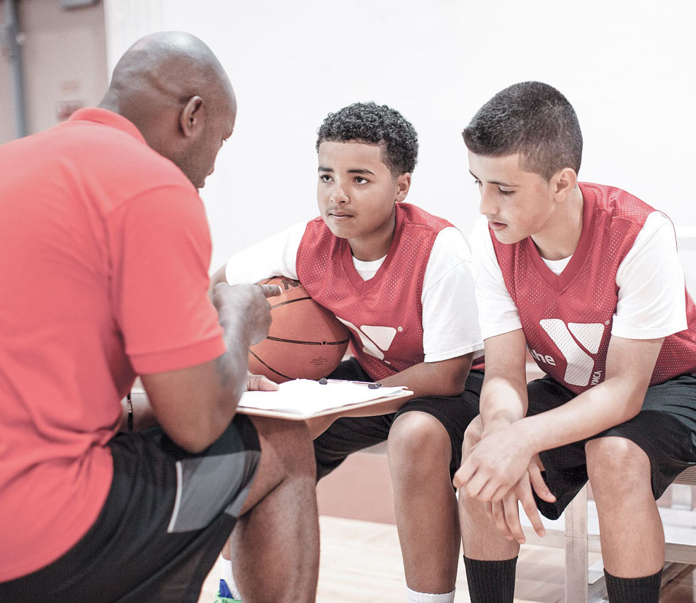 A basketball coach holds a clipboard and talks to two boys on the team