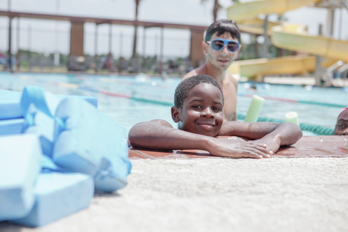 A young African American boy hangs out in the pool and rests on the pool edge, smiling