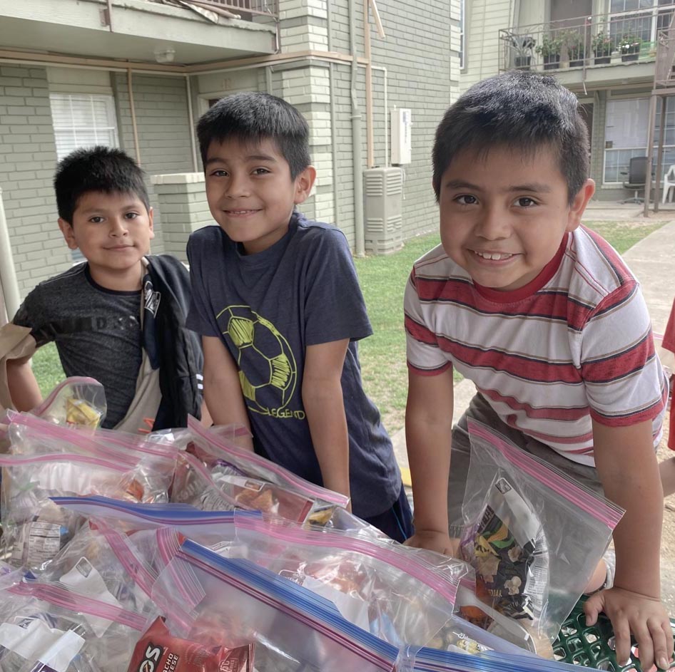 Three young boys stand at a table with packaged food ready to be distributed